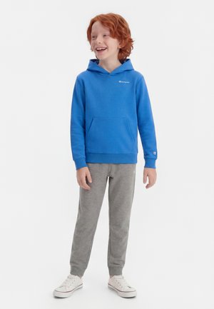 Rødhåret barn som smiler, iført blå hettegenser, grå joggebukser og hvite joggesko, stående mot en ensfarget hvit bakgrunn.