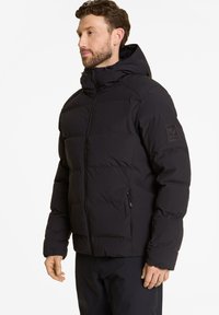Schwarze Steppjacke mit Kapuze, ausgestattet mit einem Reißverschluss an der Vorderseite, seitlichen Taschen und einem gesteppten Design. Gesticktes Logo am Ärmel.