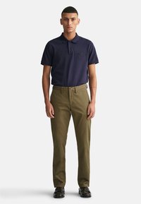 Camisa polo navy con logo bordado, combinada con pantalones chinos verde oliva y zapatos negros, de pie sobre un fondo blanco.