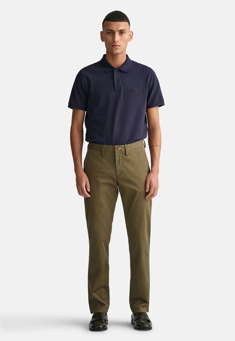 Camisa polo navy con logo bordado, combinada con pantalones chinos verde oliva y zapatos negros, de pie sobre un fondo blanco.