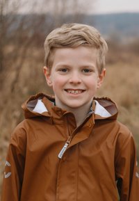 Hnědá nepromokavá bunda s kapucí, předním zipem, šedou podšívkou a bílými detaily. V pozadí jsou vysoké trávy a tlumené barvy.