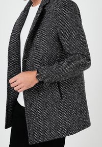 Texturovaný černobílý blazer s přiléhavým střihem, otevřeným předním dílem a bočními kapsami. Nosený přes bílou košili a doplněný hodinkami.
