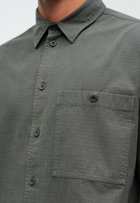 Camisa gris de botones con tejido texturizado, cuello puntiagudo y un solo bolsillo en el pecho con cierre de botón.