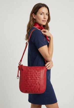 Kvinde i navyblå kjole bærer rød mønstret læder crossbody taske og rød silkehalstørklæde, ser over sin skulder mod en ensfarvet baggrund.
