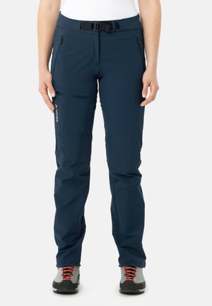 Vrouw die marineblauwe wandelbroek draagt met ritszakken, verstelbare riem en grijze wandelschoenen met oranje veters, staand met het gezicht naar voren.