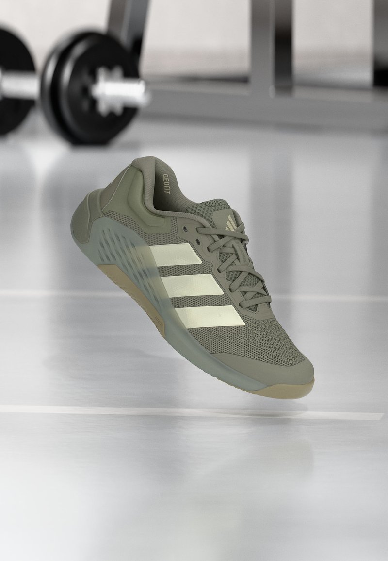Sneaker da allenamento Adidas verde oliva chiaro con tre strisce bianche, tomaia in mesh e suola strutturata, mostrata fluttuante in un ambiente da palestra.