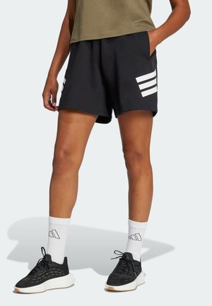 Person iført sorte atletikshorts med hvide striber, hvide crew-strømper og sorte sneakers står mod en ensfarvet baggrund.