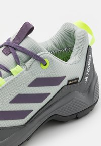 Lys grå utendørssko med mesh-overdel, lilla detaljer, neon grønne snøringer og teksturert grå såle. Har GORE-TEX merke.