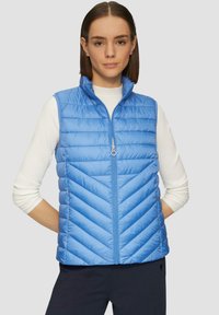 Gilet en doudoune bleu clair avec un motif en chevrons, col montant et fermeture éclair à l'avant. Porté par-dessus un haut à manches longues blanches et côtelées.