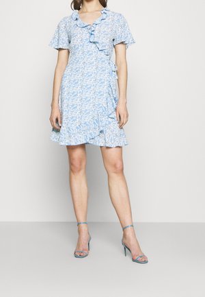 Kvinde iført en lyseblå blomsterprint slå-om-kjole med flæser, kombineret med blå højhælede sandaletter med ankelrem, stående mod en ensfarvet baggrund.