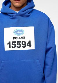 Blauwe hoodie van zachte stof met een voorzak. Opvallende witte patch met "POLIZEI" en "15594" in vetgedrukte zwarte tekst.