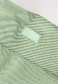 Svetlo zelena rebrasta tkanina z gladko teksturo, z vezenim obližem z napisom "self." v svetlejši odtenek zelene.