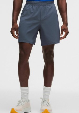 Man draagt donkergrijze sportshorts, witte kousen en lichtgekleurde hardloopschoenen, staand tegen een effen achtergrond.