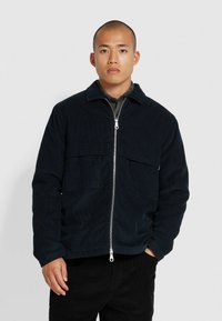 Veste en velours côtelé bleu marine avec fermeture éclair à l'avant, deux poches poitrine et un col. Présente une finition texturée et une coupe décontractée.