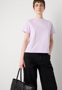 Camiseta de algodón en color lila con cuello redondo y mangas cortas. Presenta un sutil estampado de logo en el pecho. Combinada con pantalones de mezclilla negros y una bolsa de mimbre.