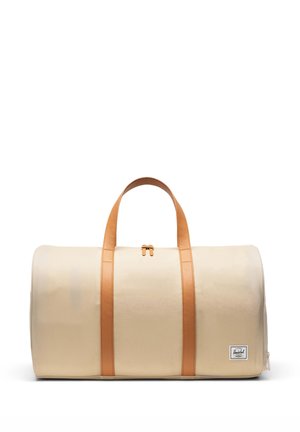 Sac polochon beige avec poignées en cuir marron clair et bandes verticales, fermeture éclair sur le dessus, et une petite écusson logo en bas à droite.