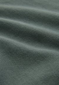 Tekstureret stof i en afdæmpet grøn farve, der viser subtile bølger og et fint strikkemønster, hvilket fremhæver blødhed og dybde.