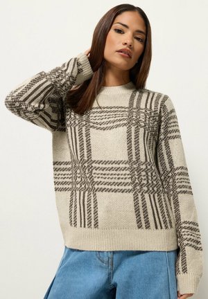 Femme portant un pull beige avec des motifs à carreaux marron et un jean bleu, posant avec une main touchant ses cheveux sur un fond uni.