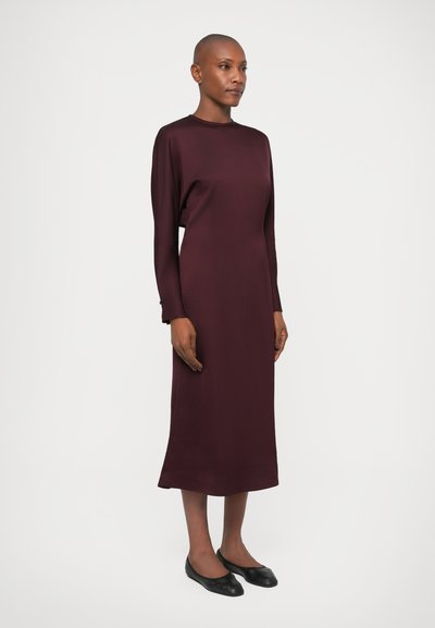Rochie burgundy cu mânecă lungă, cu un design neted și ajustat, având o siluetă dreaptă și decolteu rotund. Asortată cu pantofi negri cu talpă plată.