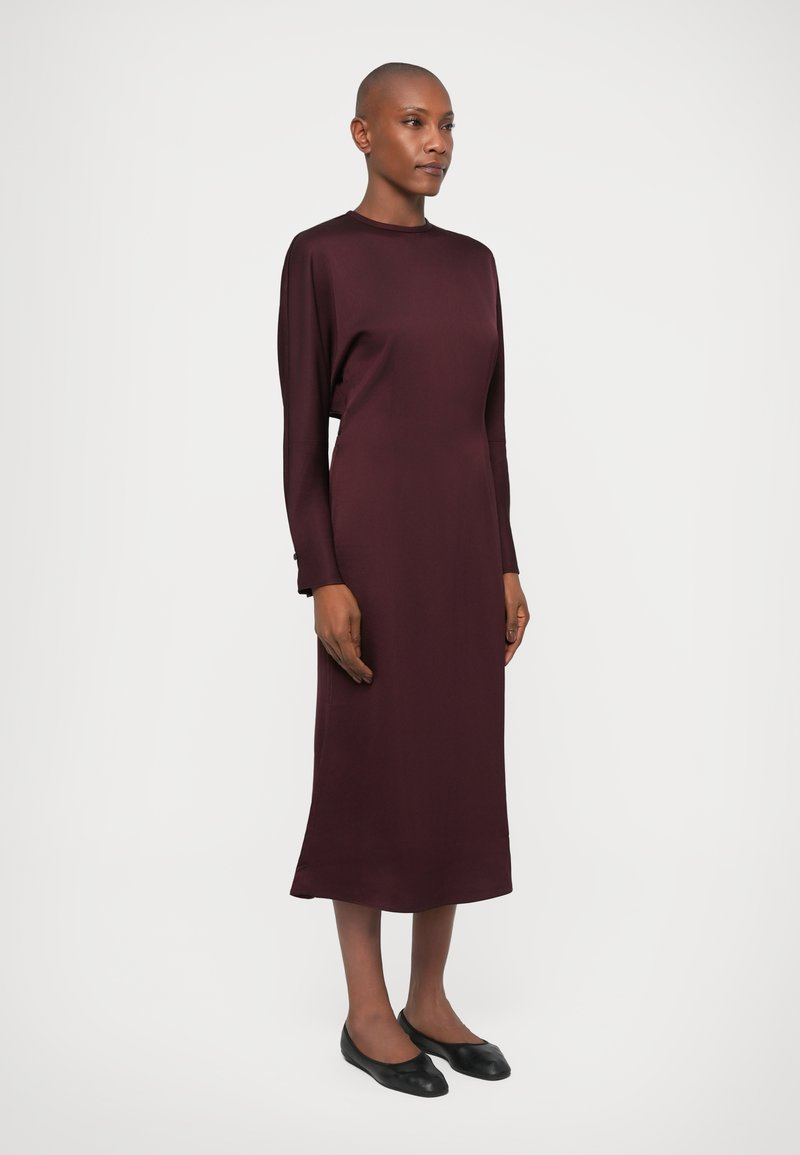 Langärmliges, burgunderfarbenes Kleid in einem glatten, taillierten Design mit gerader Silhouette und Rundhalsausschnitt. Kombiniert mit schwarzen flachen Schuhen.