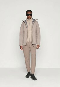 Bežs ūdensnecaurlaidīgs mētelis ar augstu apkakli, kombinēts ar bežīgu turtlneku un biksēm, valkāts ar melniem apaviem un tumšām saulessargiem.