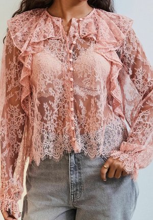 Femme portant un chemisier en dentelle florale rose avec volants, fermeture boutonnée à l'avant et manches longues, associé à un jean en denim gris taille haute.