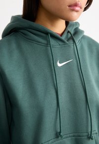 Groene hoodie van zachte stof, voorzien van een kangoeroezak en verstelbare trekkoorden. Witte Nike-logo gecentreerd op de borst.