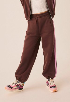 Person iført brune joggers med pinke sidelister og farverige pink og leopardmønstrede sneakers, stående med spredte ben.