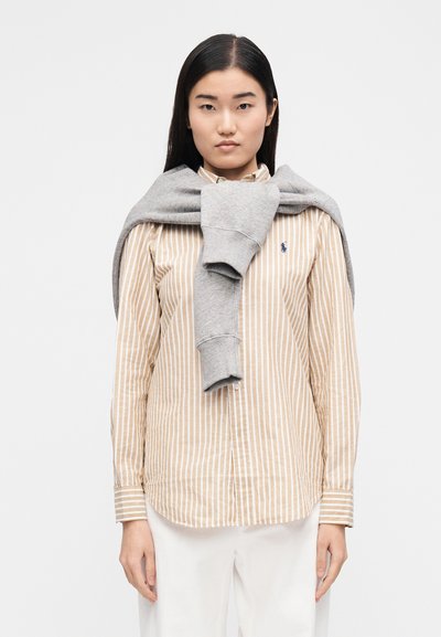 Béžovo-biele pruhované tričko so špicatým golierom, v kombinácii so sivou mikinou prehodenou cez ramená. Nosené sú biele nohavice.