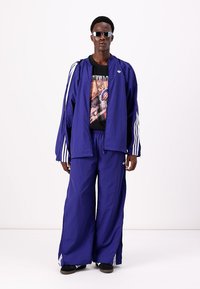 Lilla joggedress laget av lett materiale, med hvite striper langs ermene og buksene, flere lommer og en iøynefallende grafisk t-skjorte under.