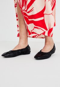 Bailarinas de cuero negro con borde elástico fruncido, que presentan un diseño de punta. Acompañadas de un vestido estampado en rojo y crema.
