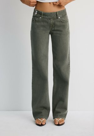 Personne portant un jean droit taille haute vert olive foncé et des sandales blanches à brides, debout devant un fond gris clair uni.