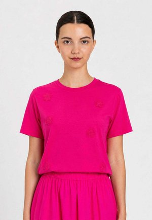 Donna con capelli scuri raccolti, indossa una maglietta rosa acceso a maniche corte con ricamo floreale e una gonna coordinata su uno sfondo semplice.