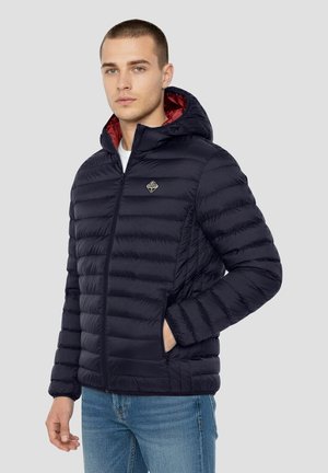 Marineblauwe donzen jas met ritssluiting aan de voorkant en capuchon, voorzien van een rood binnenvoering, gewatteerde textuur en een klein logo op de borst.