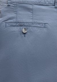 Gros plan de la poche arrière d'un pantalon bleu avec une poche passepoilée boutonnée et des détails de couture visibles.