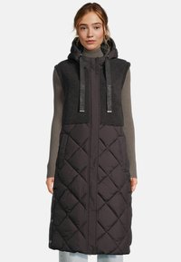 Femme portant un long manteau sans manches noir matelassé avec une capuche, une partie supérieure en polaire et un pull à col roulé côtelé gris en dessous.