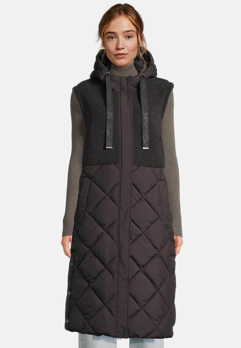 Femme portant un long manteau sans manches noir matelassé avec une capuche, une partie supérieure en polaire et un pull à col roulé côtelé gris en dessous.