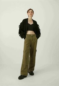 Cardigan preto com franjas sobre um topo cropped preto, combinado com calças cargo largas em verde-oliva com bolsos laterais e uma textura suave.