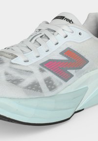 Nærbillede af hvid atletiksko med lysblå sål, hvide snørebånd og flerfarvet stribet "N"-logo på siden.