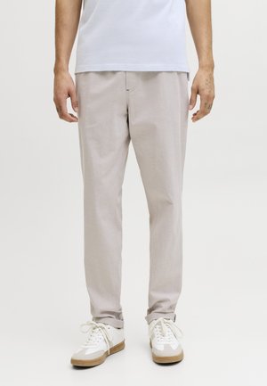 Άνδρας που φοράει ανοιχτό μπεζ παντελόνι, λευκά αθλητικά με λαστιχένιες σόλες και λευκό πουκάμισο, στέκεται μπροστά σε απλό φόντο.