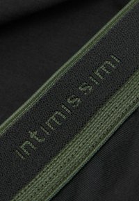 Svart tyg med en texturerad grön elastisk midjeband med upphöjd text som säger "intimisimi." Nära fokus på material och design.