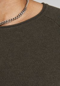 Stickad grön tröja i en texturerad tyg, med rundad halsringning och synliga sömmar. I sällskap med ett silverhalsband.