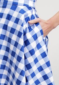 Eine Hand, die in die Tasche eines blau-weiß karierten Rocks mit Falten auf einem hellen Hintergrund gesteckt ist.