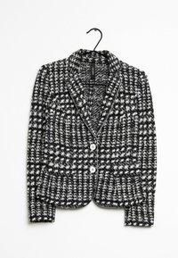 Svartvit mönstrad kavaj i texturerat tyg, med slagkrage, framknappar och rak nederkant, hängande på en krok.