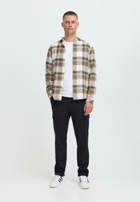 Mann står i hvite joggesko, svarte bukser, hvit t-skjorte og beige rutete skjorte med knapper, med hendene i lommene, på enkel lys bakgrunn.