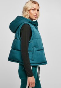 Gilet matelassé teal avec capuche et fermeture éclair à l'avant, arborant une coupe large et des poches latérales. Texture lisse avec motif matelassé et accents de cordon de serrage.