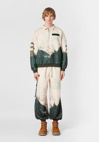 Pullover en color crema con estampado de montañas verdes y azules, con cuello con media cremallera, acompañado de pantalones baggy a juego, que presentan un estampado similar y puños elásticos.