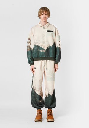 Pullover en color crema con estampado de montañas verdes y azules, con cuello con media cremallera, acompañado de pantalones baggy a juego, que presentan un estampado similar y puños elásticos.