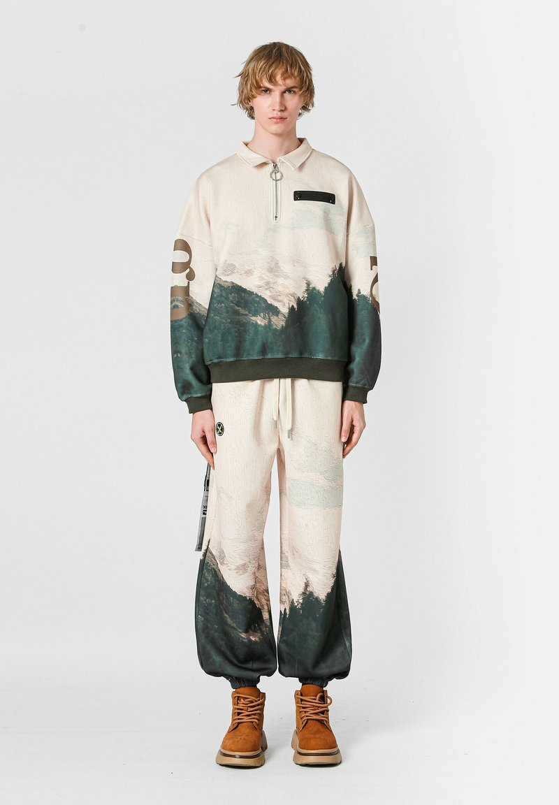 Pullover en color crema con estampado de montañas verdes y azules, con cuello con media cremallera, acompañado de pantalones baggy a juego, que presentan un estampado similar y puños elásticos.
