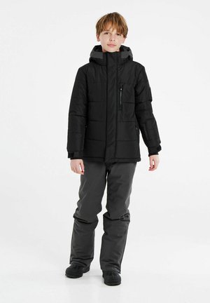 Schwarze gepolsterte Jacke mit hohem Kragen und Kapuze, ausgestattet mit Reißverschluss- und Klappentaschen. Kombiniert mit grauen isolierten Hosen und schwarzen Stiefeln.